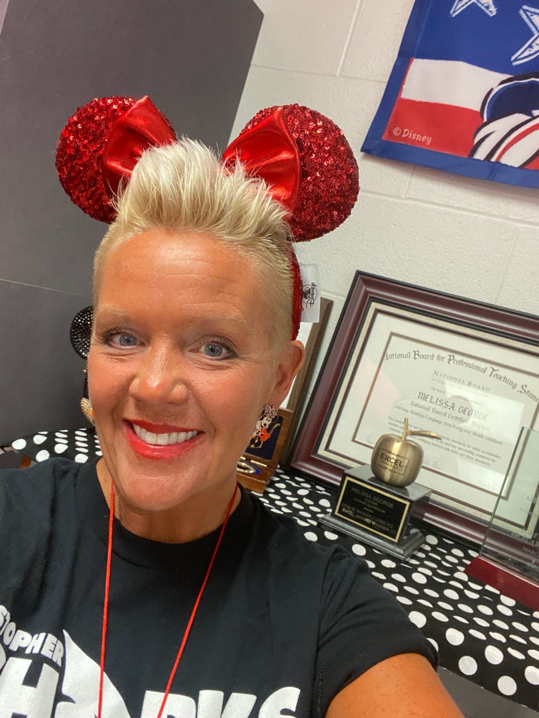 Melissa George in her classroom, smiling, in red sequin Minnie ears, with her National Board certificate and ExCEL Award apple on the shelf behind her.